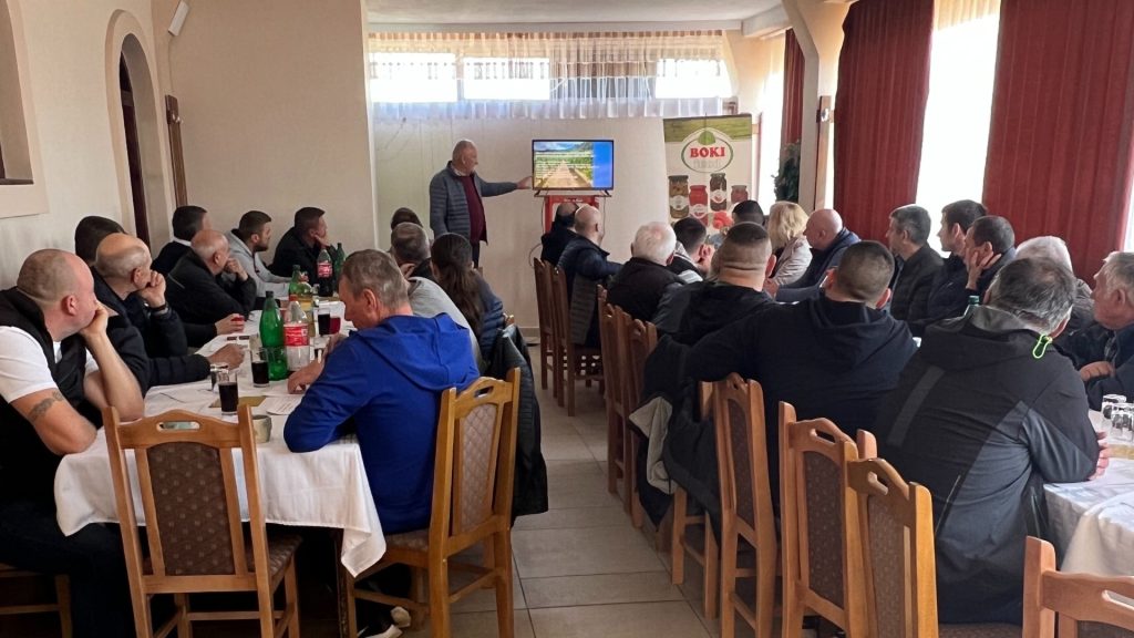 Na slici je prikazana grupa ljudi koja sedi na drvenim stolicama unutar prostorije, okrenuta ka muškarcu koji stoji i nešto prezentuje ili govori. Izgleda kao da je u toku neka vrsta sastanka ili prezentacije. U pozadini se vidi televizor na kome je prikazana prezentacija, a pored njega stoji roll-up banner sa logom i proizvodima "BOKI FOOD". Ljudi su pažljivo usmereni ka govorniku, što ukazuje na zainteresovanost za temu. Atmosfera deluje ozbiljno i fokusirano na događaj.