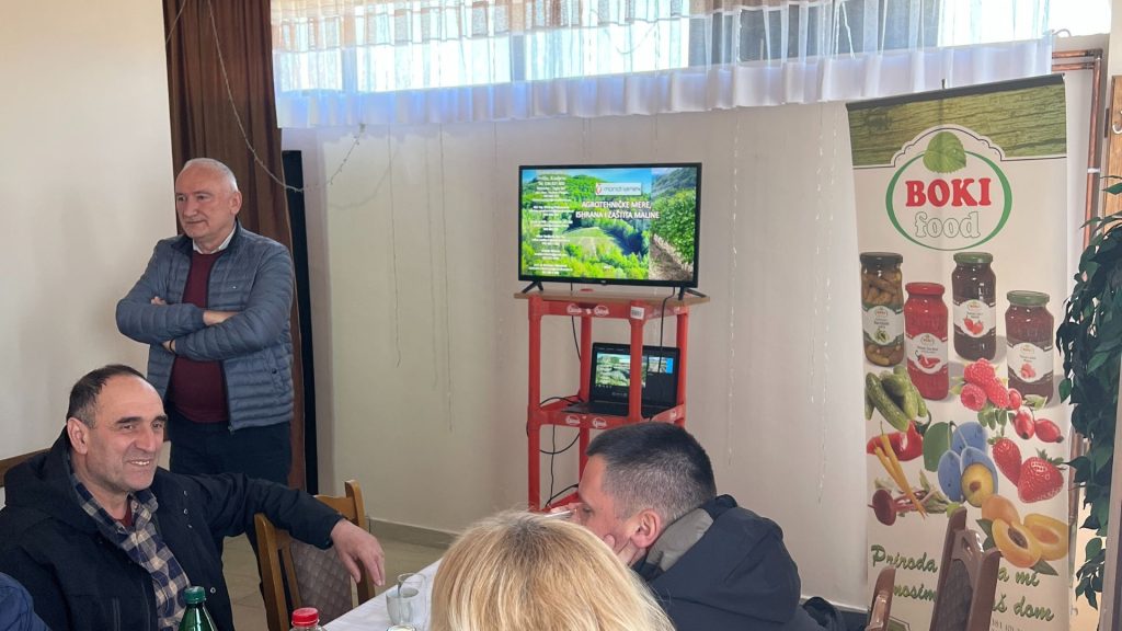 Na slici je unutrašnjost prostorije gde ljudi sede i razgovaraju. U pozadini, stariji muškarac stoji uz zid, naslonjen na štok vrata i gleda prema kameri. U centru scene je TV postavljen na crveni sto, prikazuje zeleno polje i nebo. Pored televizora je reklamni banner za "BOKI FOOD", sa slikama različitih prehrambenih proizvoda kao što su ajvar, džemovi i voće. Na prvom planu je grupa ljudi, od kojih su neki okrenuti leđima prema kameri, u opuštenoj konverzaciji. Atmosfera izgleda prijateljska i neformalna.