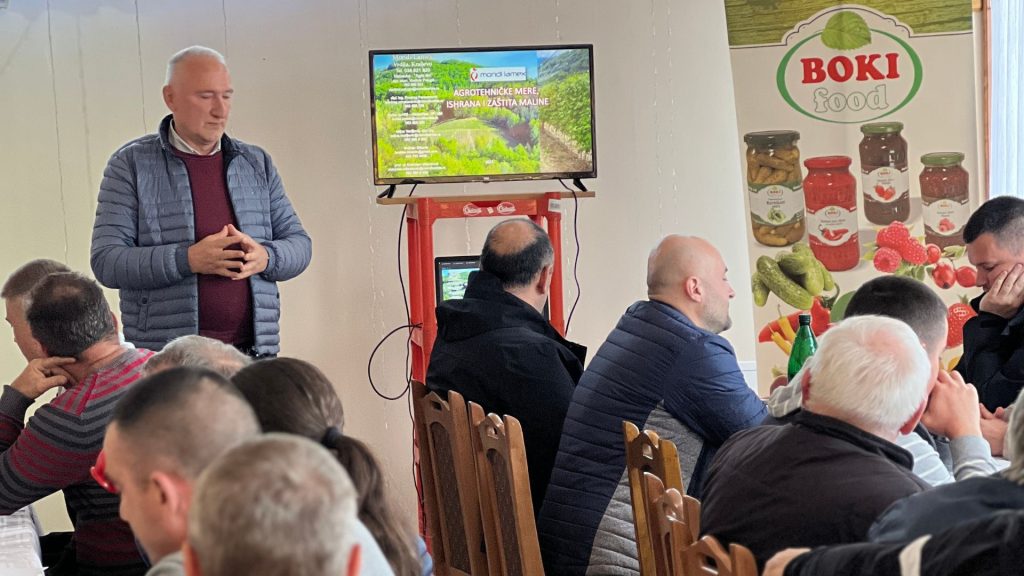 Na slici se vidi unutrašnji prostor gde grupa ljudi sedi okrenuta ka čoveku koji stoji i govori. Stoji sa prekrštenim rukama i izgleda kao da je upravo završio sa govorom ili je u pauzi dok sluša pitanje. Na televizoru u pozadini je prezentacija sa zelenim poljima i tekstom, a pored televizora nalazi se reklamni poster sa logotipom i proizvodima "BOKI FOOD". Prisutni deluju zainteresovano za prezentaciju, neki se okreću ka govorniku, dok drugi gledaju u pravcu kamere ili nešto komentarišu međusobno. Atmosfera je fokusirana ali opuštena.